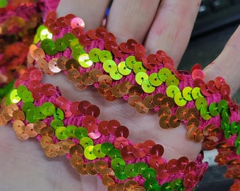 May include: Close-up of a vibrant pink sequin trim. The trim features rows of colorful sequins in shades of red, green, and gold, sewn onto a pink fabric base. This decorative trim is ideal for embellishing clothing, accessories, or craft projects.
