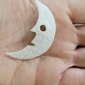 May include: A crescent moon-shaped charm with a textured, iridescent white finish and a single circular hole near the top. The object features a stylized face profile with a pointed nose and chin, crafted from a shimmering, glittery material.