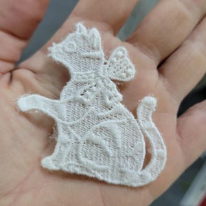 May include: A white lace cat applique with a detailed design. The cat is sitting with its tail curled and one paw raised. The applique has a decorative bow and is made of a delicate, open-weave fabric.