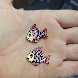 May include: Two embroidered fish patches. Each patch features a pink and purple body with a black outline, a yellow face, and red fins. The patches are small and detailed, perfect for adding to clothing or accessories.