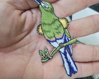 Puede incluir: Parche bordado de un colibrí posado en una rama. El ave presenta colores verde, azul y amarillo con costuras detalladas. El parche está diseñado con un contorno negro y un pequeño detalle de hoja.