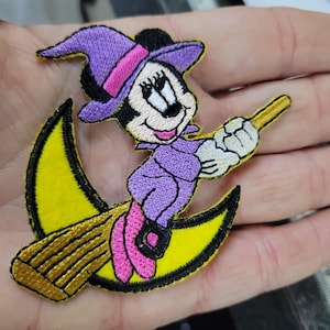 May include: Embroidered patch featuring a cartoon witch character with a purple hat and dress, riding a yellow crescent moon broom. The patch has black, yellow, pink, and white accents, and is likely for clothing or accessories.