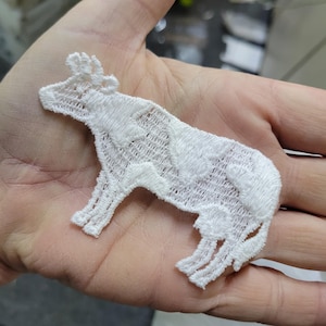 May include: A white, embroidered cow patch. The patch is made of a delicate, open-weave fabric, with the cow's form clearly defined. The cow has horns and a detailed body. The patch is held in a person's hand.