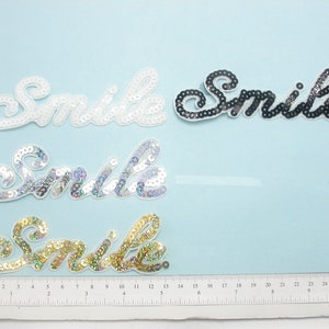 Peut inclure: Quatre patchs thermocollants avec le mot "Smile" dans différentes couleurs et styles. Les patchs sont faits de paillettes et sont disponibles en blanc, argent, noir et or.