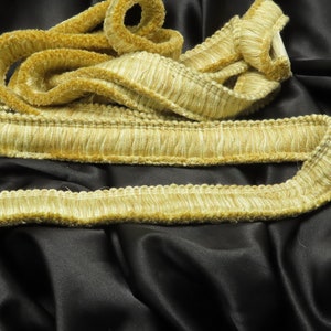 May include: A close-up of a gold-colored fringe trim with a textured surface. The trim is folded and arranged on a black fabric background.