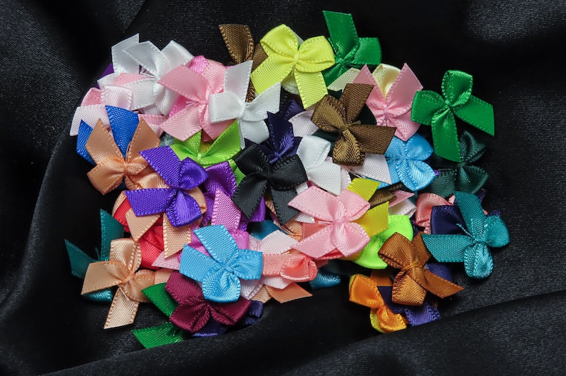 May include: A collection of colorful satin ribbon bows in various colors, including pink, blue, green, purple, and brown. The bows are arranged in a cluster on a black fabric background.