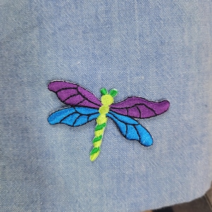 May include: A purple and blue embroidered dragonfly with a green body on a light blue fabric background.