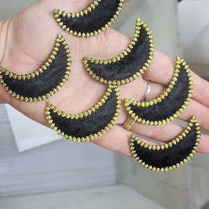 Può includere: Una collezione di sei toppe a forma di mezzaluna, ognuna caratterizzata da un centro in tessuto nero con un bordo perlato testurizzato color oro metallizzato. Queste piccole applicazioni ricamate decorative sono disposte in gruppo.