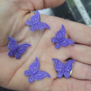 May include: Five small, purple butterfly appliques with white embroidered details. The butterflies are arranged on a light-colored surface, showcasing their delicate design. These appliques are ideal for crafting and embellishment projects.