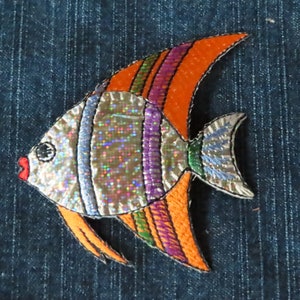 May include: A colorful embroidered fish patch with a silver iridescent body, orange fins, and purple, green, and blue stripes. The patch is on a blue denim background.