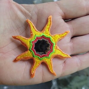 May include: A star-shaped embroidered patch featuring a central circular black mirror surrounded by concentric rings of red and green thread, with yellow and orange pointed rays extending outward in a sunburst pattern.