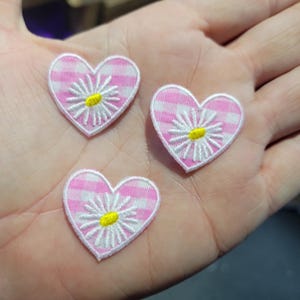 May include: Three heart-shaped patches with a pink and white gingham pattern. Each patch features a white daisy with a yellow center. The patches are embroidered with a white border.