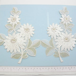 May include: Two white floral appliques with pearl accents and green leaves. The appliques are on a light blue background.