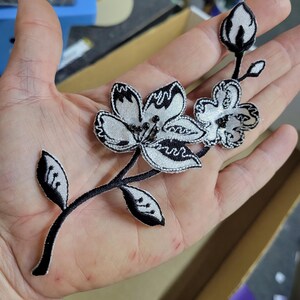 May include: A black and white floral applique featuring three flowers and two leaves. The flowers have a detailed, embroidered design with black outlines and silver-white centers. The applique is held in a hand.