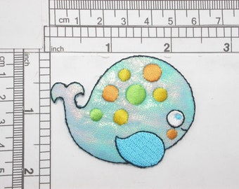 May include: A light blue whale with a yellow, orange, and green polka dot pattern. The whale has a blue tail fin and a white eye with a black pupil.