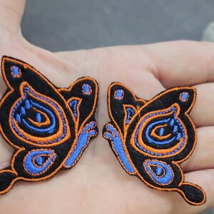 May include: Two embroidered butterfly patches featuring a black base with vibrant orange and blue thread detailing. The wings display intricate swirling patterns and small blue accents, finished with a distinct orange stitched border outline.