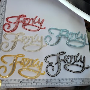 May include: Five plastic "Foxy" word charms in different colors: red, white, light blue, yellow, and black. The charms are all the same size and shape, with a cursive font.