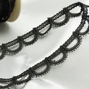 May include: Black scalloped lace trim with a decorative design. The trim is made of a delicate, woven material and is ideal for sewing and crafting projects. A roll of the trim is visible in the background.