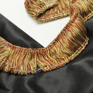 May include: A close-up of a multi-colored fringe trim with a woven texture. The fringe is made of a blend of green, yellow, and red threads. The trim is shown on a black fabric background.