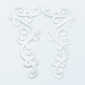 May include: Two white satin fabric embellishments with a delicate, intricate, floral design. The embellishments are shaped like a stylized letter S.