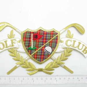 Pode incluir: Etiqueta bordada a ouro com uma faixa branca que diz "GOLF" e outra faixa branca que diz "CLUB". A etiqueta apresenta um escudo de tartã vermelho e verde com uma bandeira de golfe e uma bola de golfe.