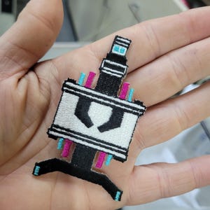May include: Embroidered patch in the shape of a microscope. The patch features a white rectangular body with black accents, and pink and blue details. The microscope has a black base and a blue top.