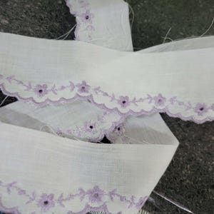May include: White fabric trim with a scalloped edge and embroidered lavender floral design. The trim is folded over itself, showing the detail of the delicate embroidery. This trim is suitable for sewing and craft projects.