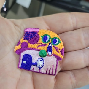 Könnte beinhalten: Gestickter Aufnäher mit einem skurrilen Pilzhaus-Design. Der Aufnäher hat einen gelben und lila Pilzhut, ein rosa und lila Dach und eine blaue Tür. Eine weiße behandschuhte Hand hält eine grüne Kugel.