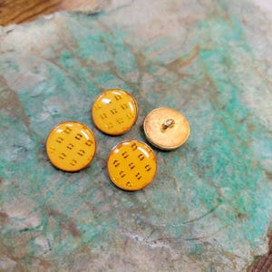 May include: Three yellow buttons with a gold geometric pattern. The buttons have a shiny finish and are perfect for sewing projects.