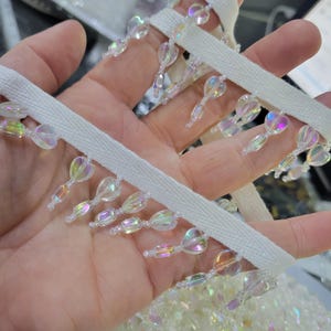 May include: Close-up of white ribbon trim with iridescent heart-shaped beads. The beads are strung along the ribbon, creating a decorative fringe. The beads reflect a rainbow of colors, adding a touch of sparkle.