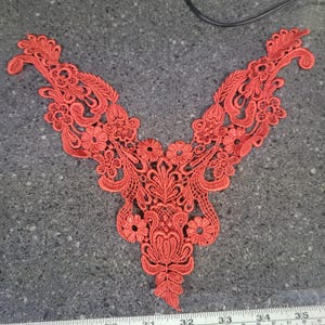 May include: A coral-colored, V-shaped lace applique with intricate floral and leaf patterns. The delicate design features symmetrical elements, including flowers and swirling details, suitable for embellishing garments or crafts.