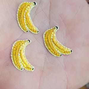 Può includere: Tre piccole toppe ricamate a forma di banana gialla. Ogni toppa presenta un design curvo con dettagliati fili giallo oro, un bordo bianco e una piccola punta nera all'estremità della banana, creando un aspetto strutturato e realistico.
