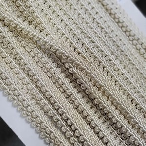 May include: A spool of off-white decorative gimp trim featuring a braided cord design with a scalloped edge. The trim is wound in multiple layers on a white cardboard backing, showcasing a consistent woven texture and a light beige color tone.