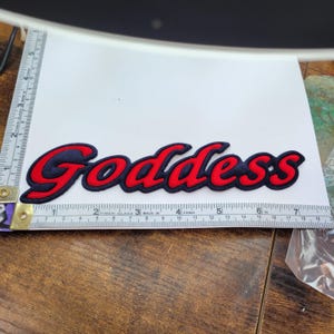 May include: Red and black embroidered patch with the word "Goddess" in a cursive font.