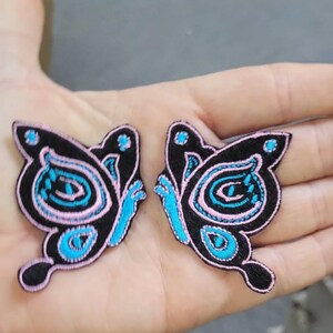 May include: Two embroidered butterfly patches featuring a black base with vibrant light blue and soft pink thread detailing. Each patch displays a symmetrical wing pattern with distinct circular and curved shapes outlined in pink and filled with blue.