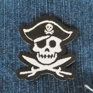 May include: A black and white pirate skull patch with a pirate hat, eye patch, and crossed swords. The skull has a white face and teeth, with a black hat featuring a skull and crossbones. The patch is on a blue denim background.
