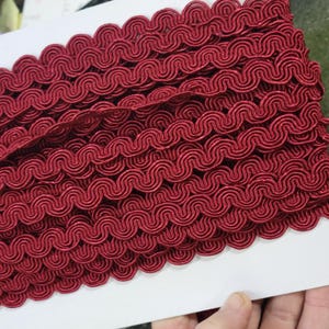 May include: A roll of burgundy decorative trim. The trim has a repeating wave pattern, creating a textured, scalloped edge. The trim is made of a soft, possibly satin-like material, and is suitable for sewing and crafting projects.