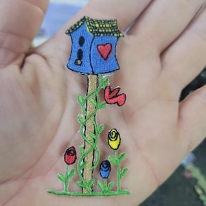 Op de afbeelding: Geborduurde patch met een blauw vogelhuisje met een rood hart, op een bruine paal met groene wijnranken en kleurrijke rozen. De patch is een levendige mix van blauw, rood, geel en groen.