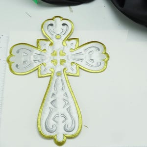 May include: A white cross with gold trim and intricate designs. The cross is made of fabric and has a raised, textured surface.
