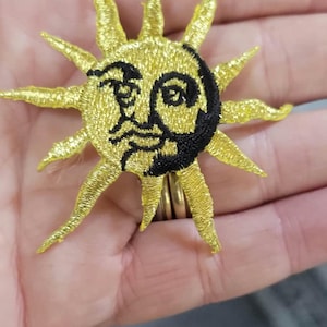 May include: A circular embroidered patch featuring a bright yellow sun with a stylised face. The sun has long, pointed rays extending outwards and a crescent-shaped black section on the right side of the face, with black thread detailing.