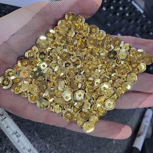 May include: Close-up of a section of black mesh fabric densely covered with gold sequins. The sequins are round, flat, and reflective, creating a shimmering effect. The arrangement forms a diamond shape. The mesh is held in a hand.