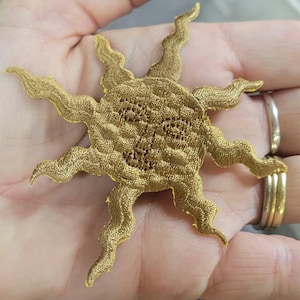 May include: A gold embroidered patch in the shape of a sun with a central face. The sun features a textured surface and multiple wavy, pointed rays extending outwards from the circular centre, creating a detailed celestial design element.