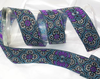 May include: Decorative ribbon featuring a black background and a repeating pattern of silver, teal, and purple floral designs. The ribbon is rolled inside a clear glass tube, highlighting its intricate details and shimmering texture. The ribbon is approximately 3.8 cm wide.