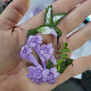 May include: Embroidered patch featuring a green and white hummingbird hovering near purple flowers. The patch has detailed stitching, showcasing the bird's wings and the floral arrangement. Ideal for adding to clothing or accessories.