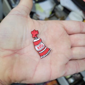 May include: A red and white embroidered patch in the shape of a tube of paint. The word "RED" is embroidered in white on the tube. The patch has a black border and a textured appearance.