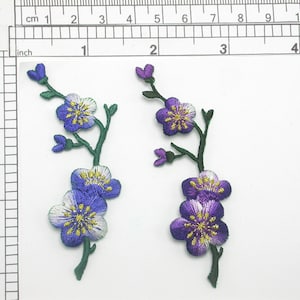 May include: Two embroidered flower appliques with green stems and leaves. The flowers are purple and blue with white centers and yellow accents.