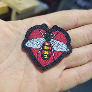 May include: Embroidered patch featuring a stylized bee design within a heart shape. The patch has a red heart background with a black border. The bee has white wings, a yellow and black body, and black details.
