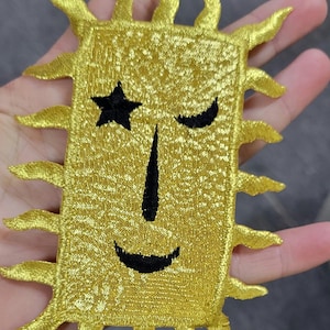 May include: A rectangular gold embroidered patch featuring a stylised sun design with radiating points along the edges. The centre displays a black embroidered face with a star-shaped eye, a closed crescent eye, a thin vertical nose, and a curved mouth.