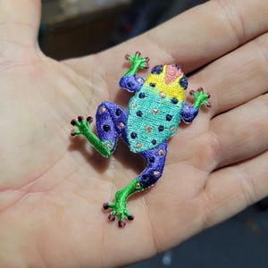 Pode incluir: Um patch de sapo bordado colorido com um design vibrante. O sapo apresenta um corpo turquesa, pernas roxas e uma cabeça amarela e rosa. O patch é embelezado com pequenas contas de cor de cobre.