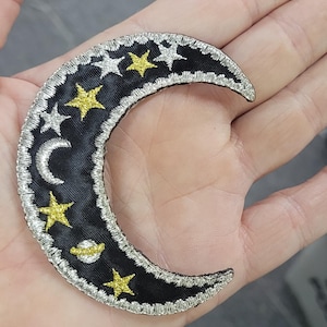 Puede incluir: Un parche bordado en forma de media luna con una base de tela negra y bordes plateados metálicos. El diseño incluye estrellas bordadas en amarillo y plata, una pequeña luna creciente plateada y un planeta plateado con un anillo.